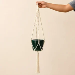 Hand holding a white macrame plant hanger with a dark green ceramic pot and a tassel at the bottom.