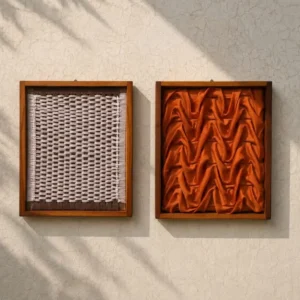 3d frame - Artoza Crafts | Where Craft Becomes Style Two framed textile swatches mounted on a textured wall: left a light woven pattern, right a folded orange fabric with chevron-like pleats.