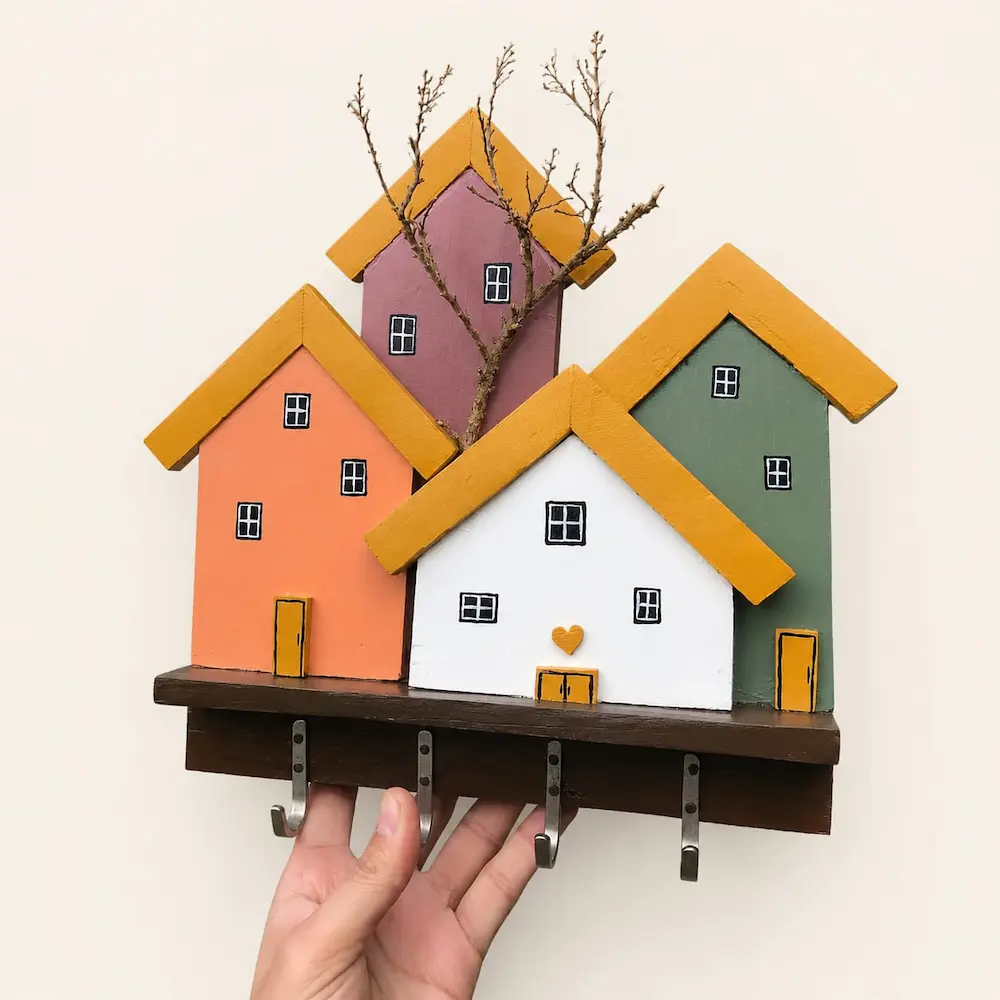 antique house keyholder - Artoza Crafts | Where Craft Becomes Style Decorative wooden wall hook rack shaped like a village of pastel-colored houses on a dark shelf, held by a hand.