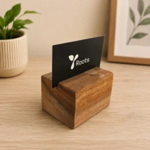 Black card with a white Roots logo inserted into a rustic wooden card holder on a light wood desk, with a potted plant and framed art in the background