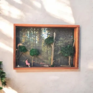 Wooden shadow box on a wall containing a miniature forest scene with trees and a small girl in a pink outfit sitting by a log.