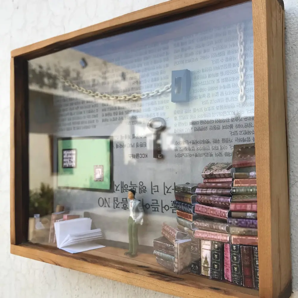 gift ideas - Artoza Crafts | Where Craft Becomes Style Miniature diorama in a glass display: tiny man near a stack of colorful books and scattered papers, with wall text in the background and a hanging chain above.