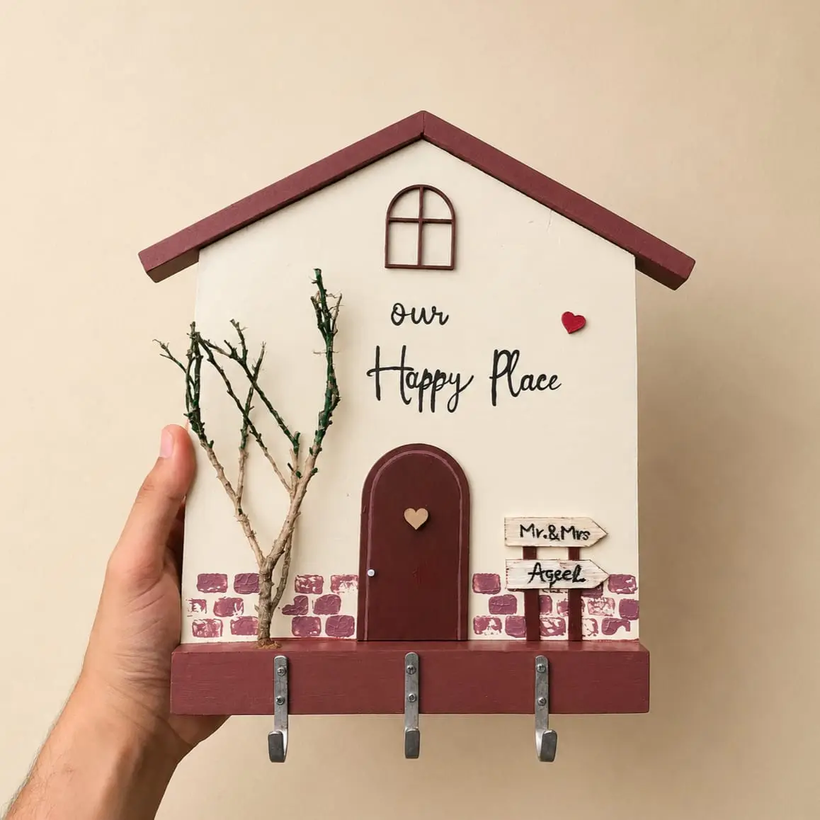 key holder - Artoza Crafts | Where Craft Becomes Style Decorative wooden house wall plaque reading 'Our Happy Place' with maroon door, twig tree, and three metal hooks at the base.