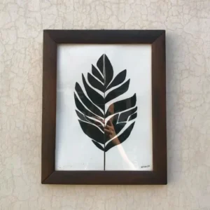 Framed black leaf silhouette artwork mounted on a light textured wall, with a wooden brown frame and a signature in the bottom right corner.