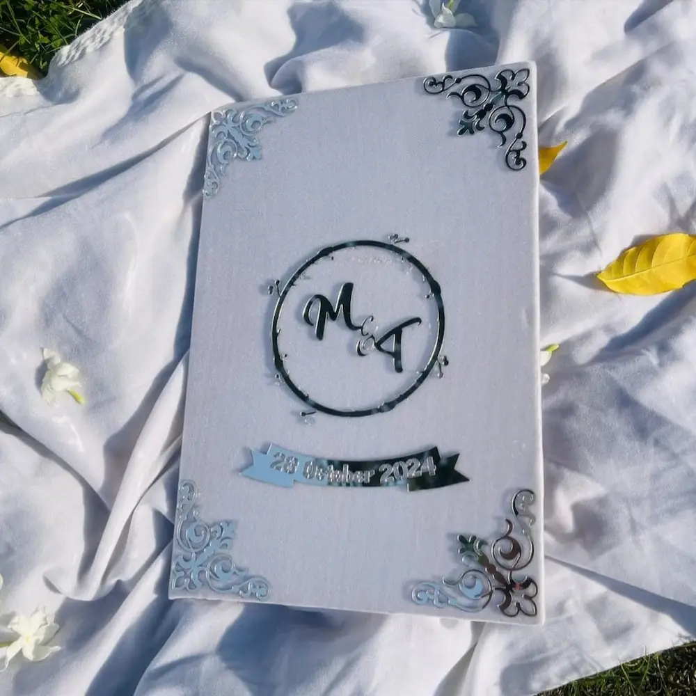 nikah booklet - Artoza Crafts | Where Craft Becomes Style White wedding album with an M A monogram inside a circular ring and a blue ribbon that reads '28 October 2024' on the cover.