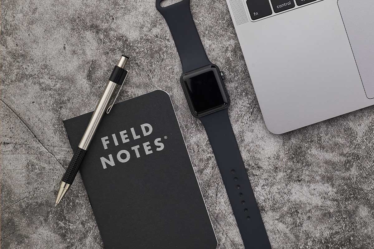 Field Notes notebook with a silver pen, black smartwatch, and a silver laptop on a textured gray surface.