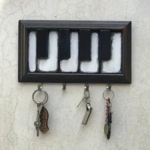 Framed decorative key rack mounted on a light wall, shaped like a piano keyboard, with three keychains hanging from the hooks below.