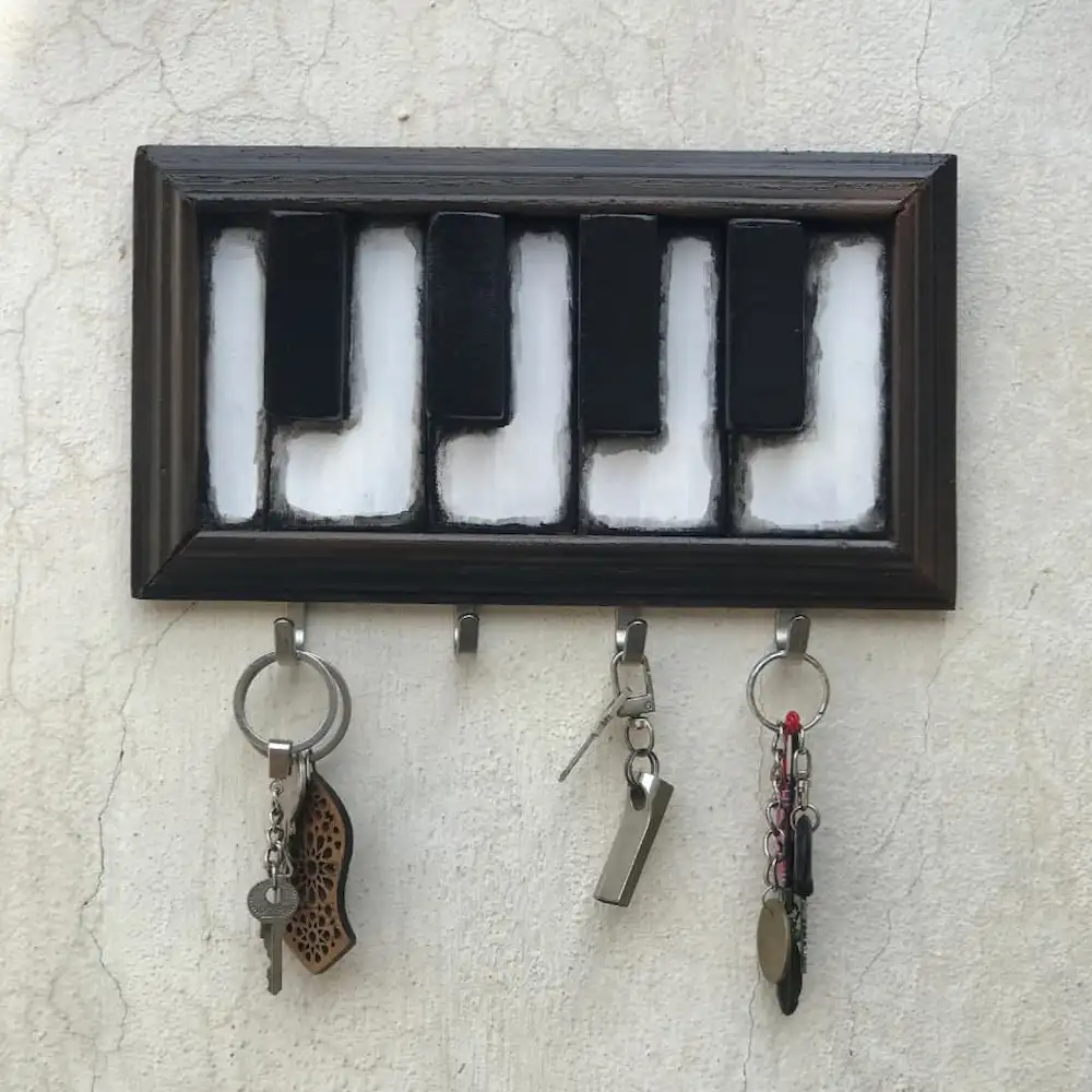 piano shape keyholder - Artoza Crafts | Where Craft Becomes Style Framed decorative key rack mounted on a light wall, shaped like a piano keyboard, with three keychains hanging from the hooks below.