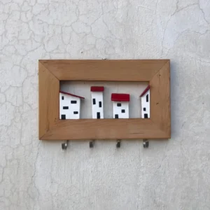 Wooden frame holding a row of four white miniature houses with black windows and red roofs, mounted on a textured wall, with four hooks beneath the frame.