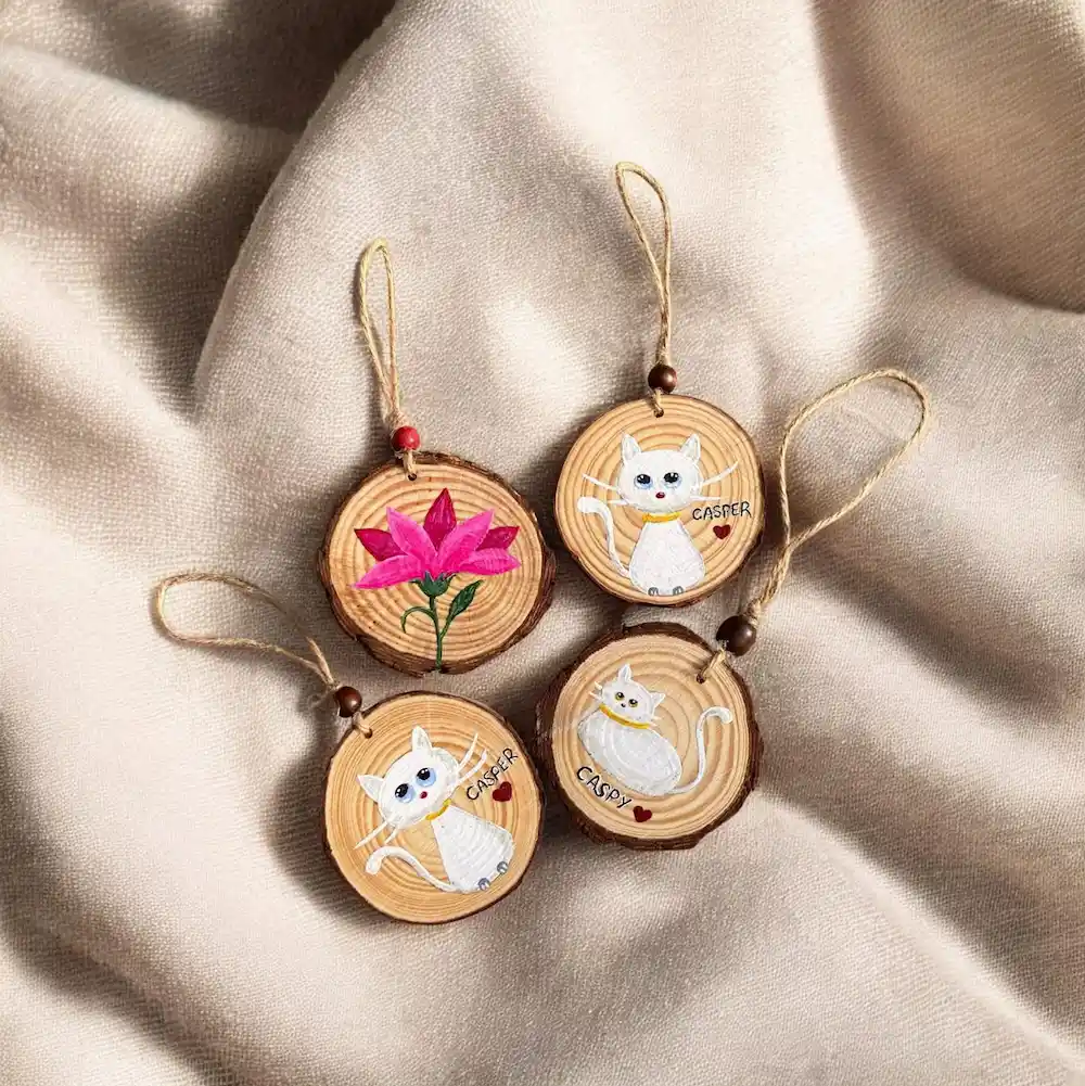 wooden slice customise keychains - Artoza Crafts | Where Craft Becomes Style Four round wooden ornaments with hand-painted white cats and a pink flower, hung on twine.